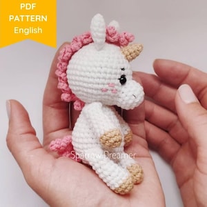 Könnte beinhalten: Ein weißes gehäkeltes Einhorn mit rosa und beigefarbenen Akzenten. Das Einhorn hat eine rosa Mähne und einen rosa Schwanz, beige Hufe und schwarze Augen. Das Einhorn wird in den Händen einer Person gehalten.