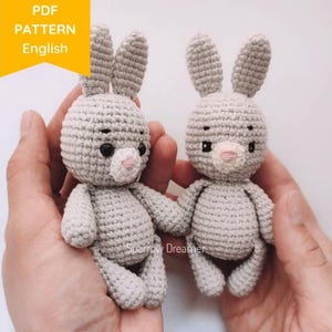 Peut inclure: Deux petits lapins crochetés gris avec des yeux noirs et des nez roses sont tenus dans des mains. Les lapins ont de longues oreilles et sont faits avec un point serré. L'image comprend le texte "PDF PATTERN English" dans une bannière jaune et le texte "Sparrow Dreamer".