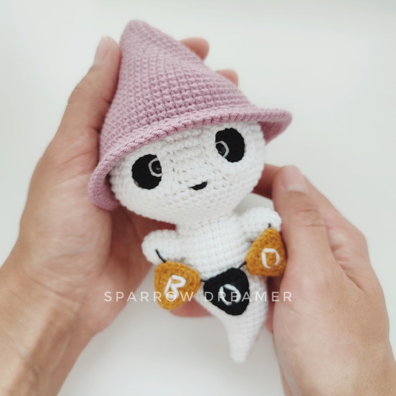 May include: A white crocheted ghost wearing a pink crocheted hat and holding a banner that says "BOO".