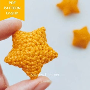 Puede incluir: Primer plano de una estrella de ganchillo en un tono amarillo vibrante. La estrella se sostiene entre los dedos, mostrando su superficie texturizada y puntadas detalladas. La imagen incluye el texto "PDF PATTERN English" en una pancarta amarilla. Estrellas adicionales borrosas en el fondo.