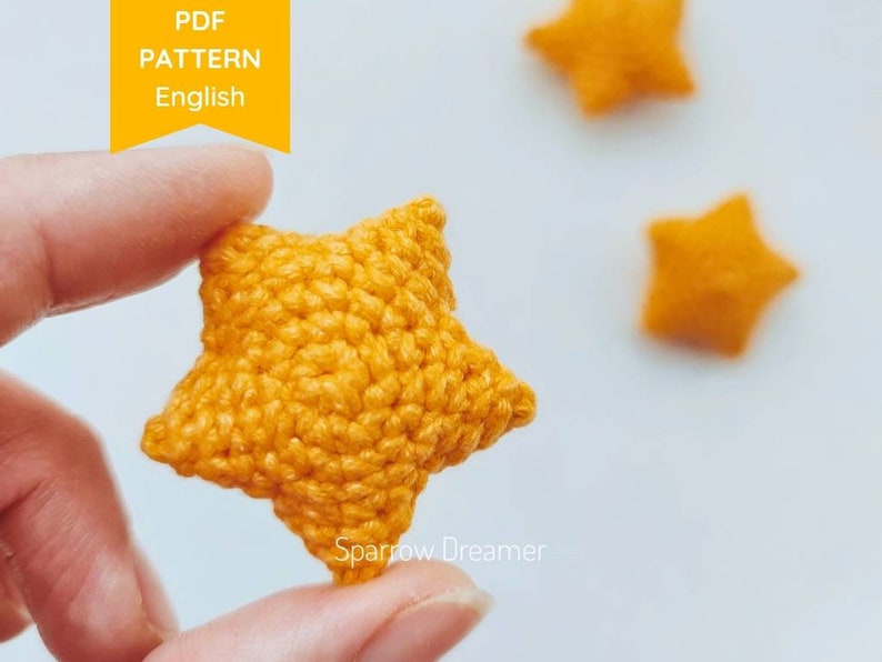 Puede incluir: Primer plano de una estrella de ganchillo en un tono amarillo vibrante. La estrella se sostiene entre los dedos, mostrando su superficie texturizada y puntadas detalladas. La imagen incluye el texto "PDF PATTERN English" en una pancarta amarilla. Estrellas adicionales borrosas en el fondo.