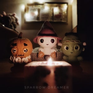 May include: Three crocheted Halloween decorations: a pumpkin, a ghost wearing a witch hat, and a green monster. The decorations are lit by a candle in the center of the image.