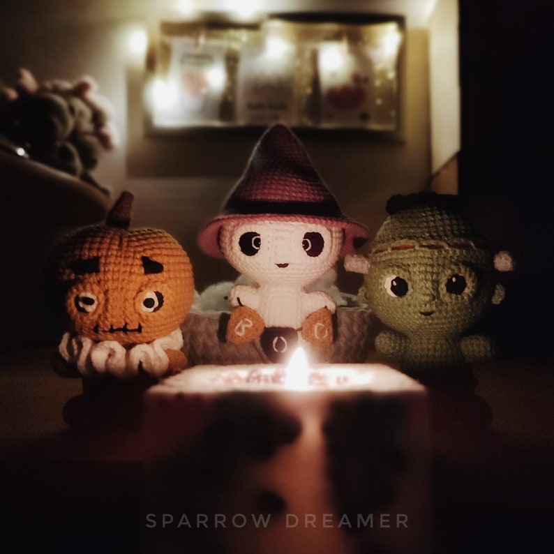May include: Three crocheted Halloween decorations: a pumpkin, a ghost wearing a witch hat, and a green monster. The decorations are lit by a candle in the center of the image.