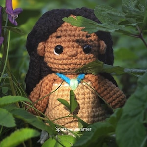 May include: A handmade crochet doll with black hair, brown skin, and a tan dress. The doll has large black eyes and a blue and green necklace. The doll is surrounded by green leaves and purple flowers. The doll is a handmade item.