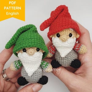 以下が含まれることがあります： 赤い帽子をかぶったグノームと緑の帽子をかぶったグノームの2体の編みぐるみ。両方のグノームは白いひげ、灰色の体、黒い靴をしています。グノームは人の手に持たれています。