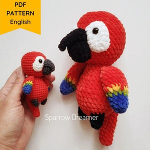 May include: Two red crochet parrot plushies, one held in a hand. The parrots have black beaks, white eye patches, and colourful wings. The larger parrot is standing upright. A yellow banner with the text "PDF PATTERN English" is in the upper left corner.