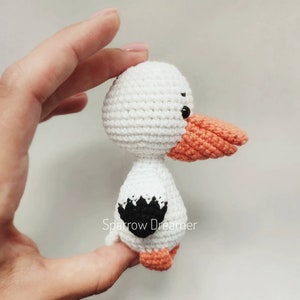 Op de afbeelding: Een handgemaakte witte gehaakte vogel met een oranje snavel en poten, en zwarte accenten. De vogel wordt in een hand gehouden, wat het gedetailleerde vakmanschap van het amigurumi-ontwerp laat zien. De vogel heeft een zwart-wit ontwerp op zijn rug.