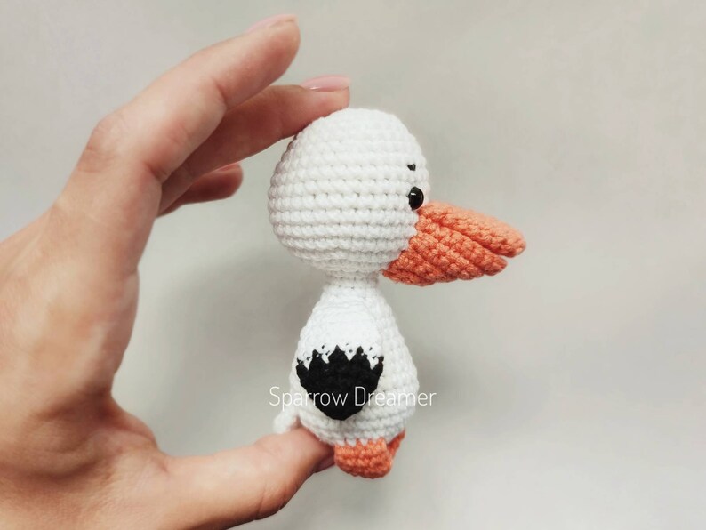 Op de afbeelding: Een handgemaakte witte gehaakte vogel met een oranje snavel en poten, en zwarte accenten. De vogel wordt in een hand gehouden, wat het gedetailleerde vakmanschap van het amigurumi-ontwerp laat zien. De vogel heeft een zwart-wit ontwerp op zijn rug.