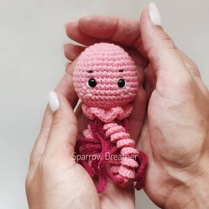 Op de afbeelding: Een handgemaakte, roze gehaakte kwal met donkerroze tentakels en zwarte ogen. De kwal wordt in open handen gehouden tegen een lichte achtergrond. De tekst "Sparrow Dreamer" is zichtbaar.