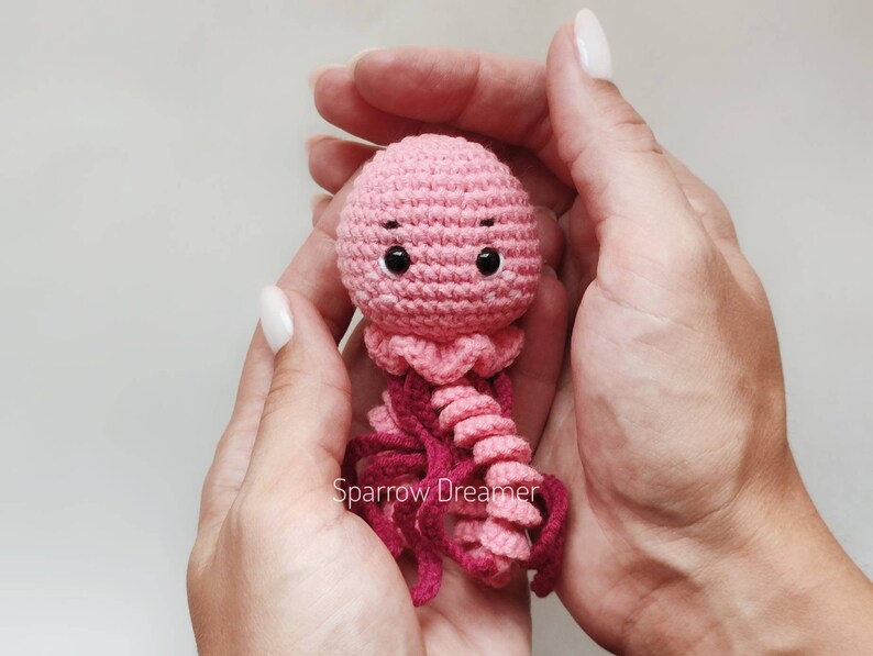 Op de afbeelding: Een handgemaakte, roze gehaakte kwal met donkerroze tentakels en zwarte ogen. De kwal wordt in open handen gehouden tegen een lichte achtergrond. De tekst "Sparrow Dreamer" is zichtbaar.