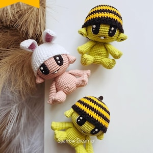 May include: Three handmade crochet dolls in the image. One is pink with a white bunny ear hat, and two are yellow with black and yellow striped hats. The dolls have large black eyes and are lying down. The image includes the text "Crochet PATTERN PDF in Eng".
