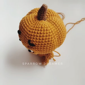 May include: A crocheted pumpkin with a brown stem and a cute face. The pumpkin is orange and has a white string attached to it.