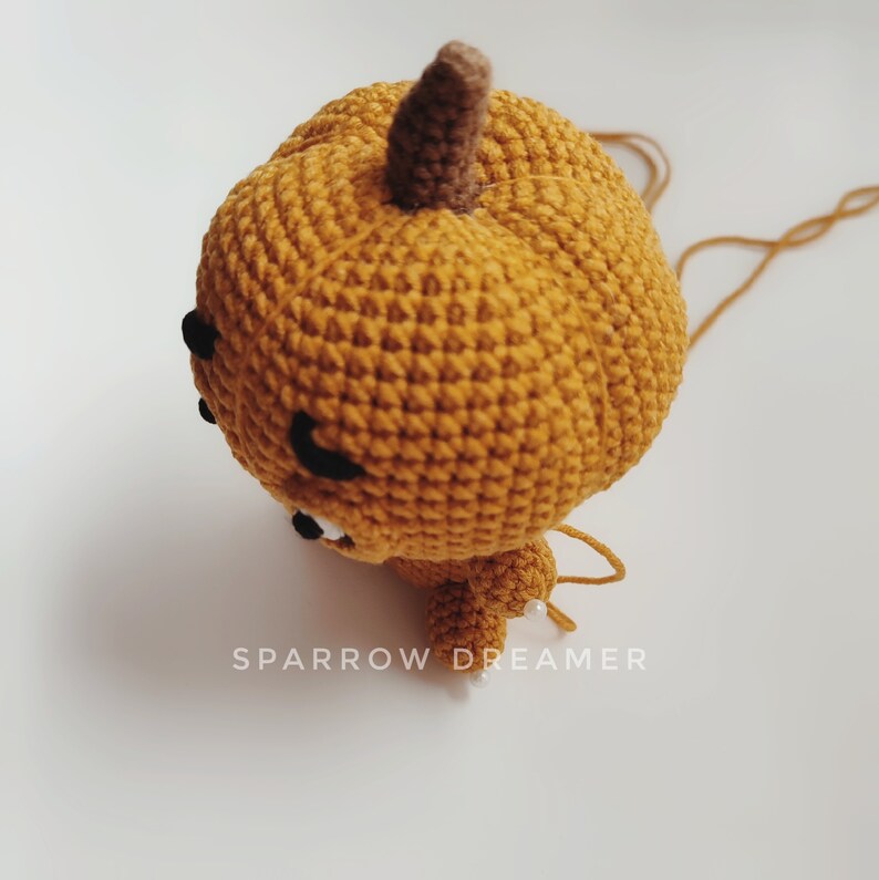 May include: A crocheted pumpkin with a brown stem and a cute face. The pumpkin is orange and has a white string attached to it.