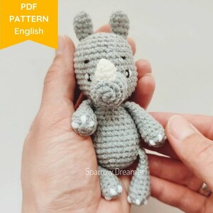 May include: A gray crocheted rhinoceros amigurumi toy with white details on the horn and feet. The toy is being held in a person's hands.