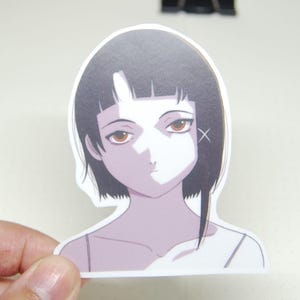 Può includere: Un adesivo con un'illustrazione in stile anime di una ragazza con capelli corti neri e un top bianco. L'adesivo è tenuto sopra un quaderno aperto con un nastro blu. Altri oggetti sono visibili sullo sfondo.