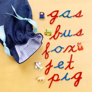 May include: A denim bag with light blue ribbons, alongside colorful toy figures and wooden cursive letters spelling "gas", "bus", "fox", "jet", and "pig". The toys include a gas pump, school bus, fox, airplane, and pig.