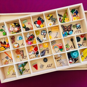 May include: A wooden tray with multiple compartments filled with colourful decorative pins and charms. The tray is divided into sections, each containing a variety of small objects, including a butterfly, a car, and a watermelon slice. A yellow zip is on the side.