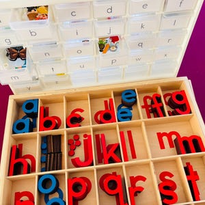 May include: Wooden alphabet learning set with red and blue letters in a divided wooden box. The top section has small clear plastic containers with letters and small objects. The letters are in a variety of fonts.