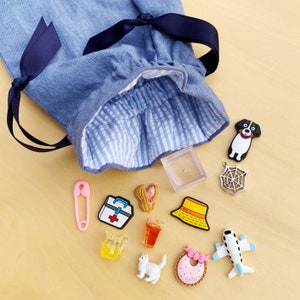 May include: A blue denim drawstring bag with a navy ribbon, open to show various charms. Charms include a dog, spiderweb, safety pin, first aid kit, peanut, drink, cat, baby bib, airplane, and a yellow hat.