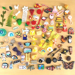 May include: A selection of small, decorative embellishments in various colours, including a red car, a yellow school bus, a butterfly, a guitar, and a zipper. Other items include a ring, a question mark, and a slice of watermelon.