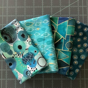 May include: Four folded fabric squares in shades of teal, turquoise, and navy blue. Each square features a unique pattern, including abstract circles, leaf shapes, geometric designs, and dotted patterns. Gold accents are visible in some designs.