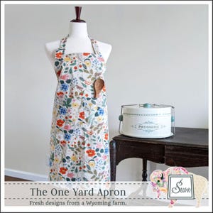 May include: A floral print apron on a mannequin with a wooden spoon in the pocket. The apron has a white background with red, blue, and yellow flowers. A white cake carrier sits on a dark wooden table.