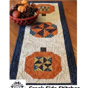 May include: A quilted table runner titled Pumpkin Patch measuring 15 inches by 41 inches, featuring three orange pumpkin motifs with patterned centers on a cream background, bordered by dark blue fabric, with a red bowl of pinecones nearby.