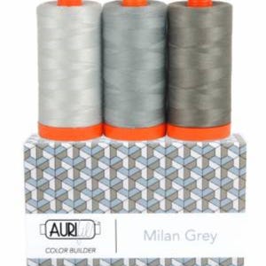 May include: Three spools of Aurifil Milan Grey thread in varying shades of gray, with orange tops, sit atop a box with a geometric pattern. The box features the Aurifil logo and the text "Milan Grey Color Builder."