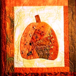 以下が含まれることがあります： 様々な秋の色彩と質感でパッチワークされたかぼちゃが描かれたキルトの壁掛け。かぼちゃは白地に囲まれ、茶色とオレンジの縁取りが施されています。