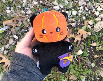 May include: A black plush cat with a pumpkin face and a purple trick-or-treat bag. The cat has white eyes and a stitched orange mouth. The bag has a yellow crescent moon.