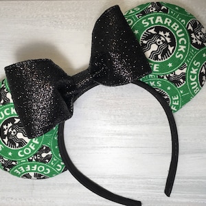 Op de afbeelding: Een zwarte glitter strik haarband met twee groene en witte Starbucks logo oren. De haarband is zwart en de oren zijn cirkelvormig. Het Starbucks logo is op de oren gedrukt.