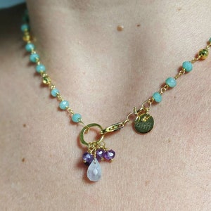 May include: Gold-toned necklace featuring light blue beads and gold accents. The necklace has a pendant with purple and clear gemstones. A small gold charm with text is attached near the clasp. The necklace is designed to be worn around the neck.
