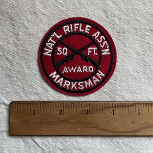 May include: A red and black embroidered patch with the text "NAT'L RIFLE ASS'N 50 FT. AWARD MARKSMAN". The patch features a crossed rifle design.