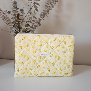 May include: A quilted, rectangular pouch with a yellow floral pattern on a light yellow background. The pouch has a gold zipper and a small white label that reads "Sarah Logan Designs". Dried eucalyptus branches are in the background.