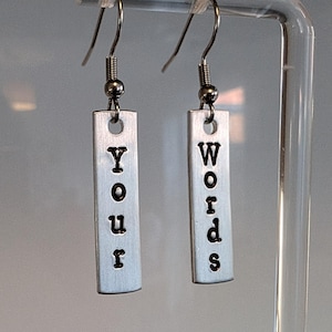 May include: A pair of silver-coloured dangle earrings. Each earring has a rectangular metal tag with the words "Your" and "Words" stamped in black, lowercase letters. The earrings hang from silver-coloured fish hook ear wires.