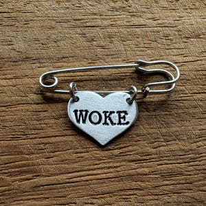 Könnte beinhalten: Sicherheitsnadel in Silber mit herzförmigem Anhänger. Der Anhänger trägt das Wort „WOKE“ in schwarzen Großbuchstaben. Die Nadel liegt auf einer strukturierten, braunen Holzoberfläche.