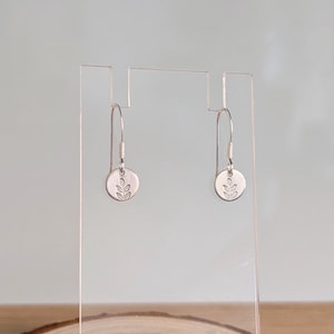 May include: A pair of silver-toned drop earrings featuring small circular charms. Each charm is engraved with a simple leaf motif. The earrings are suspended from thin, curved metal hooks that attach to the top of the circular pendants.