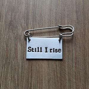 Może przedstawiać: Srebrna agrafka z prostokątną metalową zawieszką. Na zawieszce wytłoczono słowa „Still I rise” pogrubioną czcionką. Agrafka jest umieszczona na drewnianym tle.