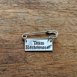 May include: A silver-colored safety pin with a rectangular tag that reads "Team Herbivore" in a bold, sans-serif font. A small leaf graphic is to the right of the text. The pin is set against a textured, brown wooden surface.