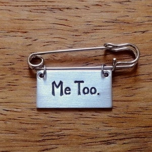 May include: A silver safety pin with a small rectangular tag attached. The tag is engraved with the words "Me Too."