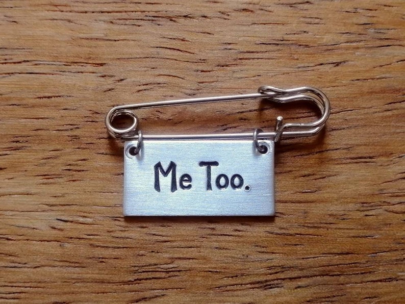 May include: A silver safety pin with a small rectangular tag attached. The tag is engraved with the words "Me Too."