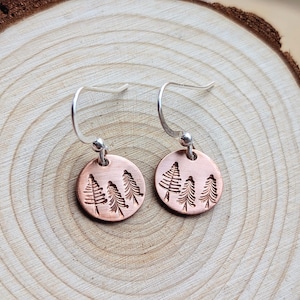 Peut inclure: Une paire de boucles d'oreilles pendantes circulaires de couleur cuivre, ornées d'un motif estampé représentant trois conifères sur chaque disque. Les boucles d'oreilles sont fixées à des crochets en métal argenté pour un rendu simple et net.