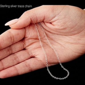 May include: A delicate sterling silver trace chain is displayed against a dark background. The chain is held in an open hand, showcasing its fine links and subtle shine. The text "Sterling silver trace chain" is visible.