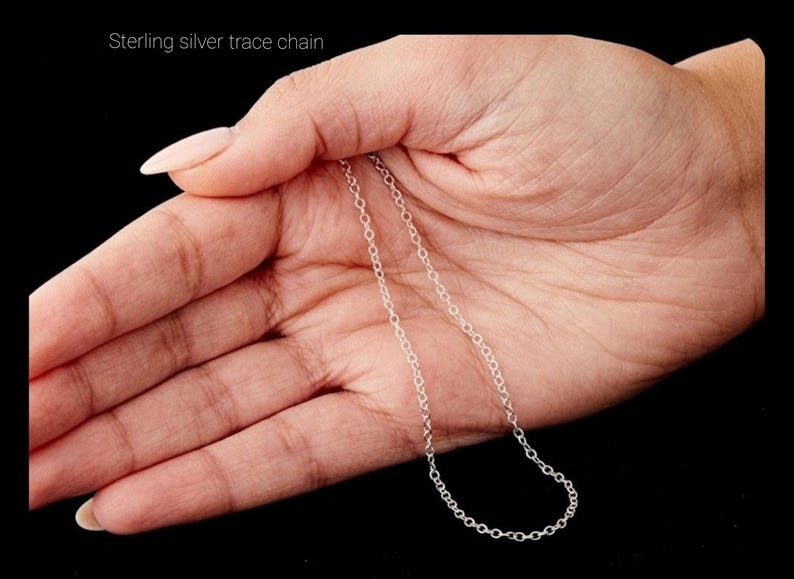 May include: A delicate sterling silver trace chain is displayed against a dark background. The chain is held in an open hand, showcasing its fine links and subtle shine. The text "Sterling silver trace chain" is visible.