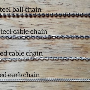 May include: Four different types of metal chains are displayed against a wooden background. The chains include stainless steel ball chain, stainless steel cable chain, silver plated cable chain, and silver plated curb chain. Each chain has a clasp.