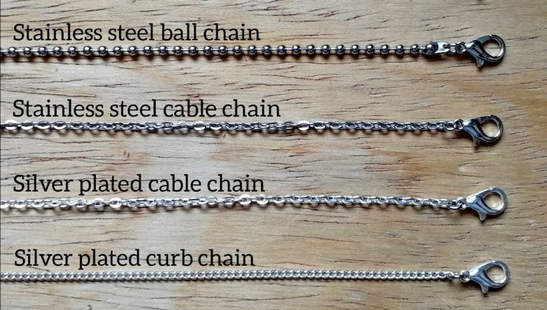 May include: Four different types of metal chains are displayed against a wooden background. The chains include stainless steel ball chain, stainless steel cable chain, silver plated cable chain, and silver plated curb chain. Each chain has a clasp.