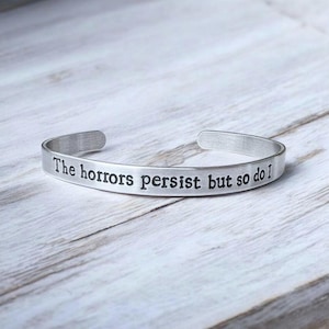 Puede incluir: Una pulsera de puño plateada con la frase grabada "The horrors persist but so do I". La pulsera se muestra sobre una superficie de madera clara. El metal tiene un acabado cepillado.