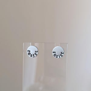 Op de afbeelding: Een paar zilverkleurige oorstekers. Elke oorbel is een kleine, cirkelvormige schijf met het woord "resist" in zwarte kleine letters. De oorbellen worden gepresenteerd op een transparante standaard tegen een neutrale achtergrond.