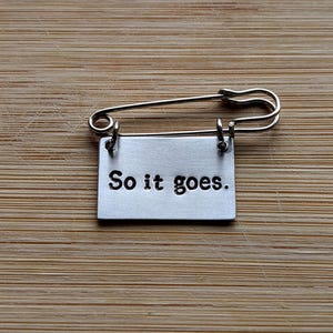 Może przedstawiać: Srebrna agrafka z prostokątną metalową zawieszką. Na zawieszce wytłoczono napis "So it goes." Agrafka leży na drewnianej powierzchni z jasnobrązowym usłojeniem.