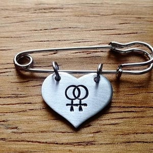 Puede incluir: Un imperdible de metal plateado con un dije en forma de corazón sujeto por dos pequeñas argollas. El dije de corazón tiene un acabado cepillado y está grabado con un símbolo negro que consiste en dos signos femeninos entrelazados en el centro.